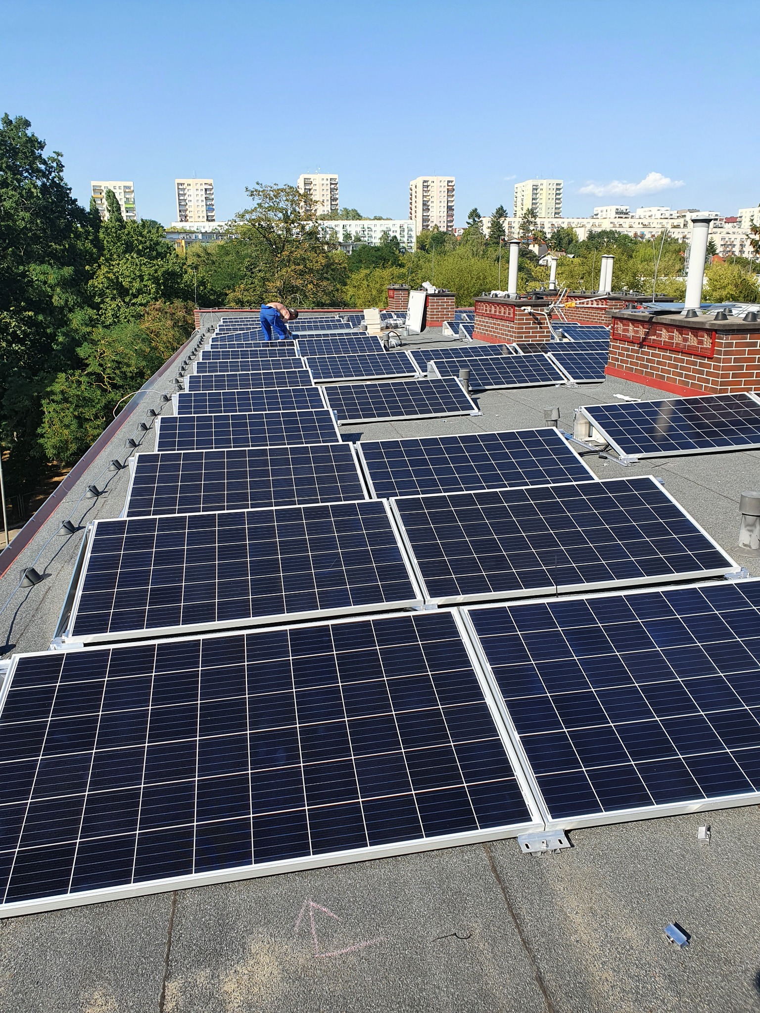 Montaż paneli słonecznych na dachu budynku z ceglanymi kominami, w tle zieleń drzew i budynki mieszkalne w słoneczny dzień.