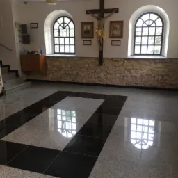 Wnętrze odrestaurowanego budynku w Warszawie z kamienną ścianą, dwoma łukowymi oknami z widokiem na zewnątrz, krucyfiksem i portretami na ścianie oraz nową, błyszczącą podłogą z ciemnymi akcentami.