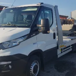 Biała laweta Iveco z platformą transportową, zaparkowana na placu budowlanym z reklamą materiałów budowlanych w tle.