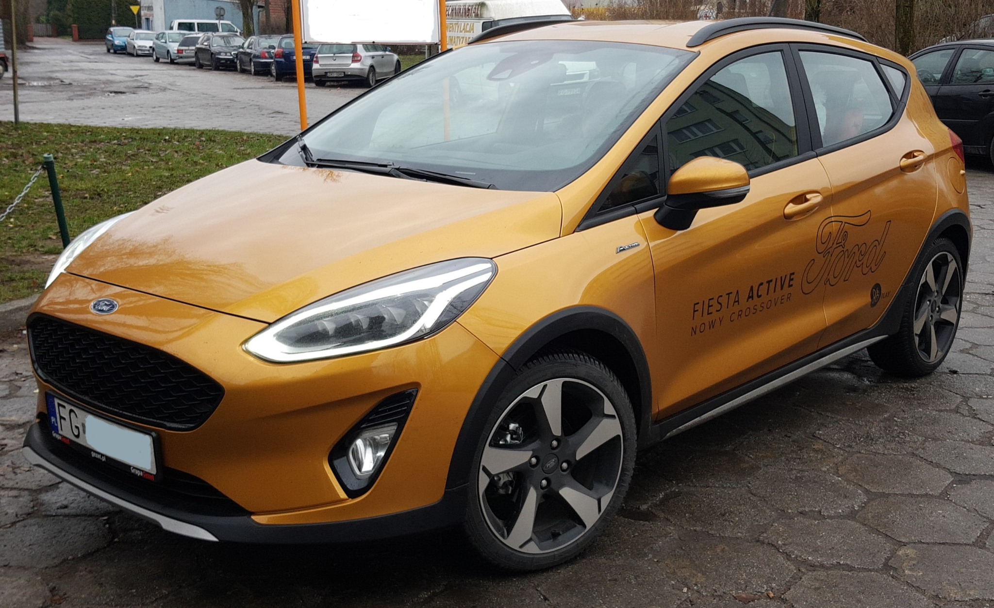 Złoty Ford Fiesta Active, nowy crossover, zaparkowany na brukowanej ulicy, widok z przodu i boku, z widocznym logo Forda.