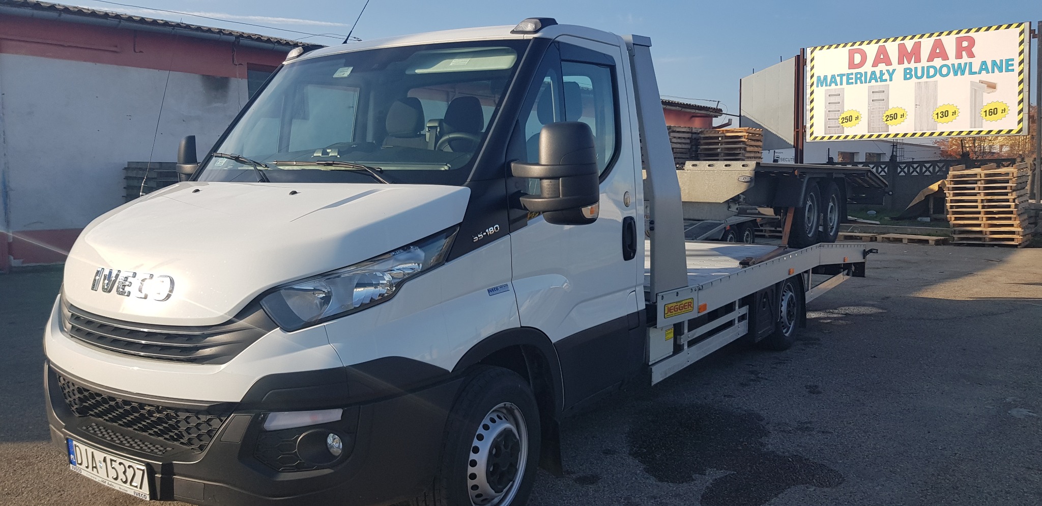 Biała laweta Iveco z platformą transportową, zaparkowana na placu budowlanym z reklamą materiałów budowlanych w tle.