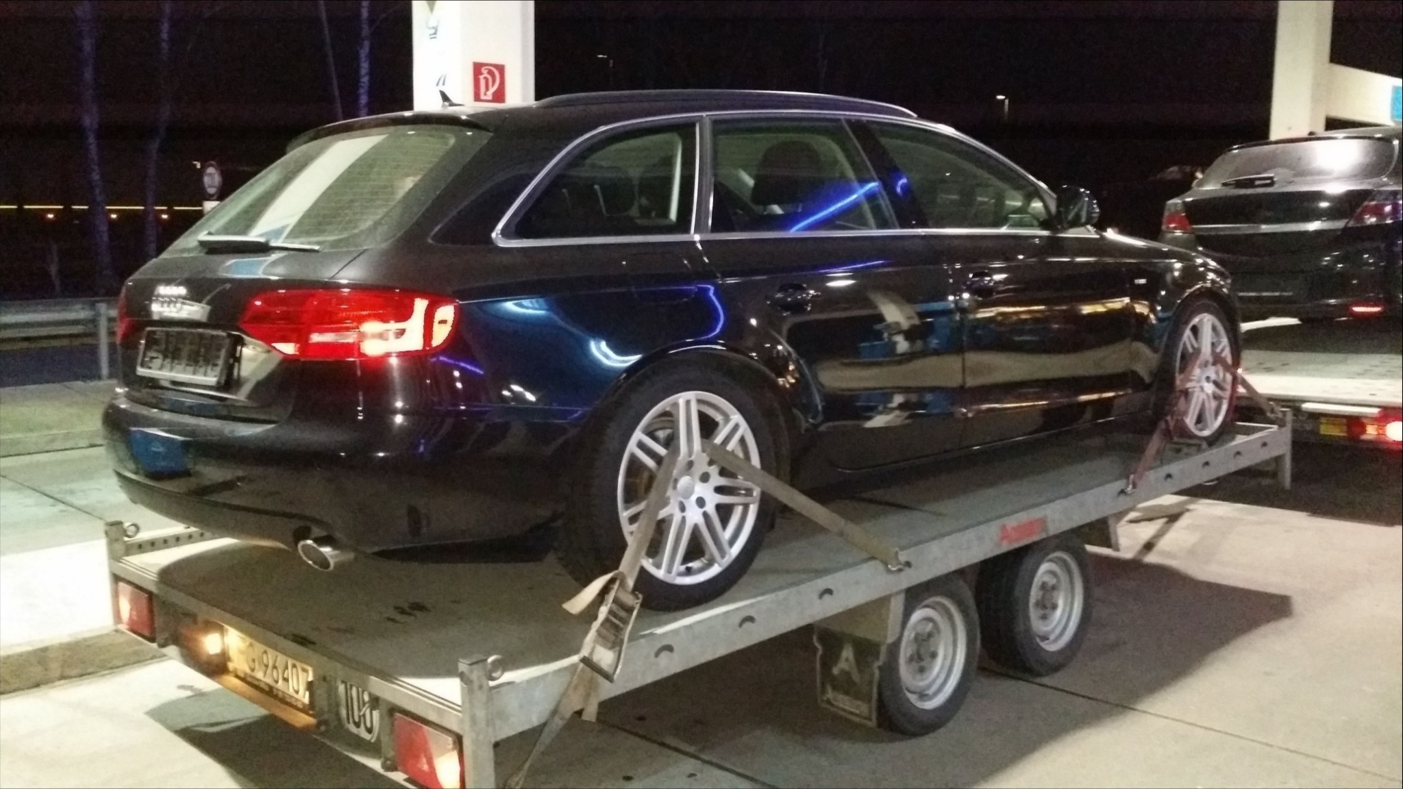 Czarny samochód kombi marki Audi na platformie lawety zabezpieczony pasami transportowymi, widoczne światła tylne i felgi aluminiowe.