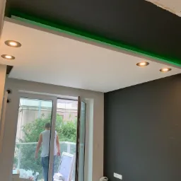 Nowoczesne wnętrze z podwieszanym sufitem z oświetleniem LED i punktowymi lampami, widok na balkon przez otwarte drzwi balkonowe, ściana pomalowana na ciemny grafit.