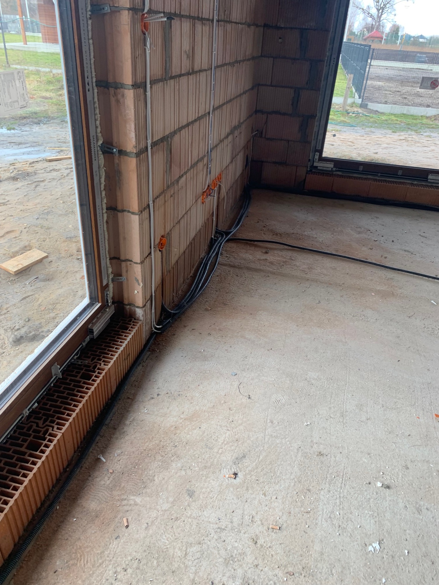 Wnętrze w stanie surowym z widocznymi przewodami elektrycznymi poprowadzonymi wzdłuż ściany z cegły ceramicznej, okno z zamontowanymi listwami i uszczelnieniami, oraz peszel przy podłodze.