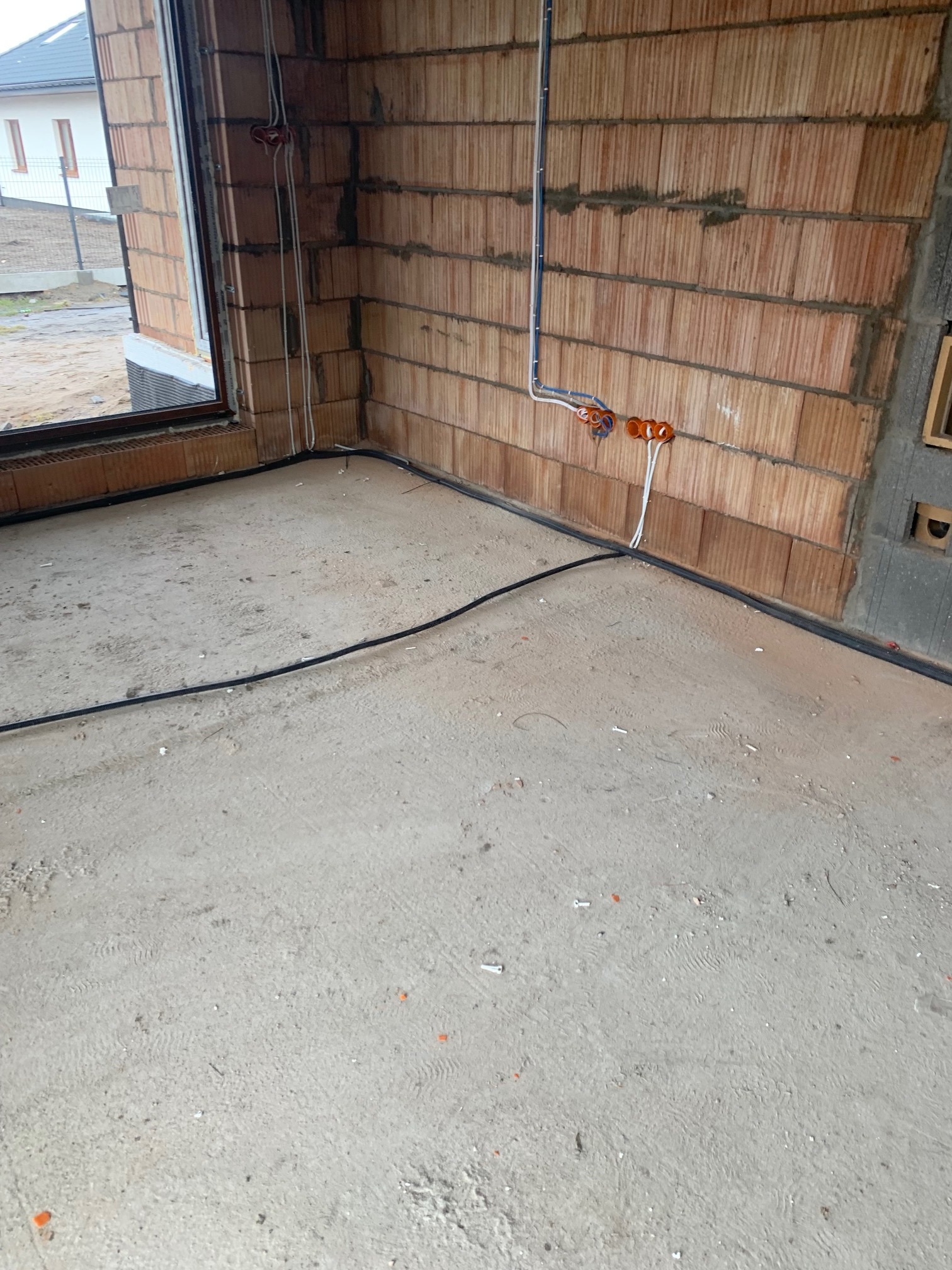 Surowy stan wnętrza z widocznymi przewodami elektrycznymi prowadzonymi po ścianach z cegły dziurawki oraz wzdłuż podłogi, przygotowane pod montaż instalacji.