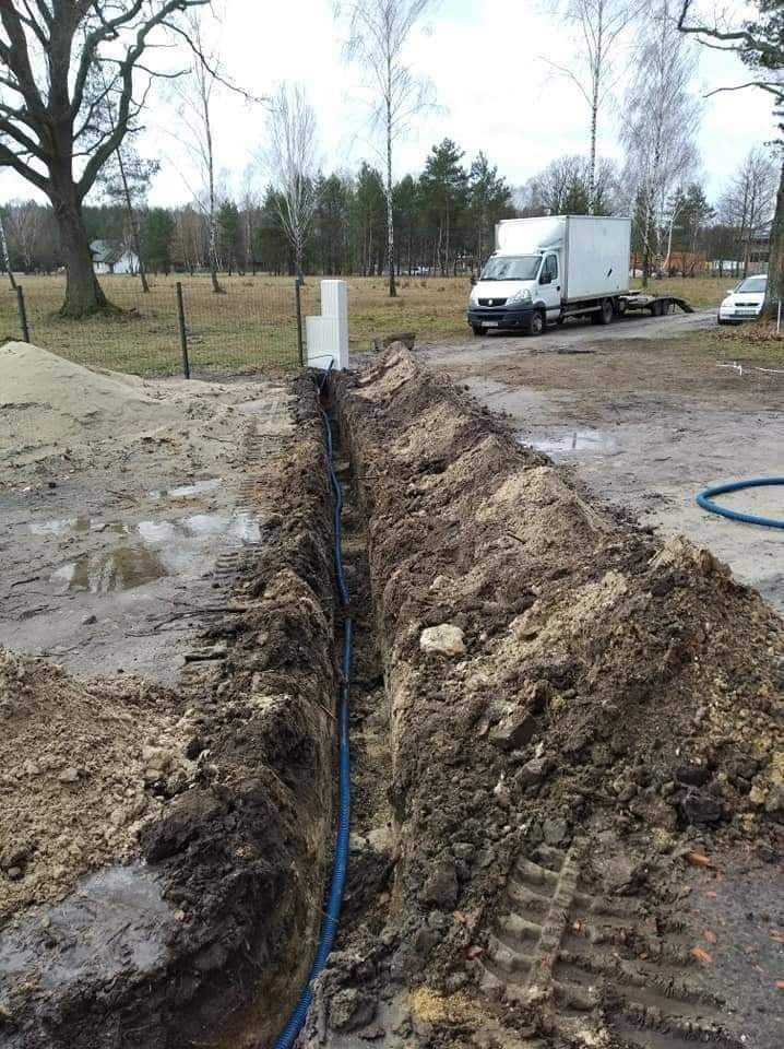 Wykonany wykop w ziemi z ułożonym niebieskim kablem w karbowanej osłonie, widoczne białe pustaki i samochód dostawczy w tle.