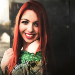 Portret uśmiechniętej kobiety z długimi, rudymi włosami, w kraciastej bluzce z zieloną, marszczoną górą, na rozmytym tle miejskim z elementami architektury i refleksami światła.