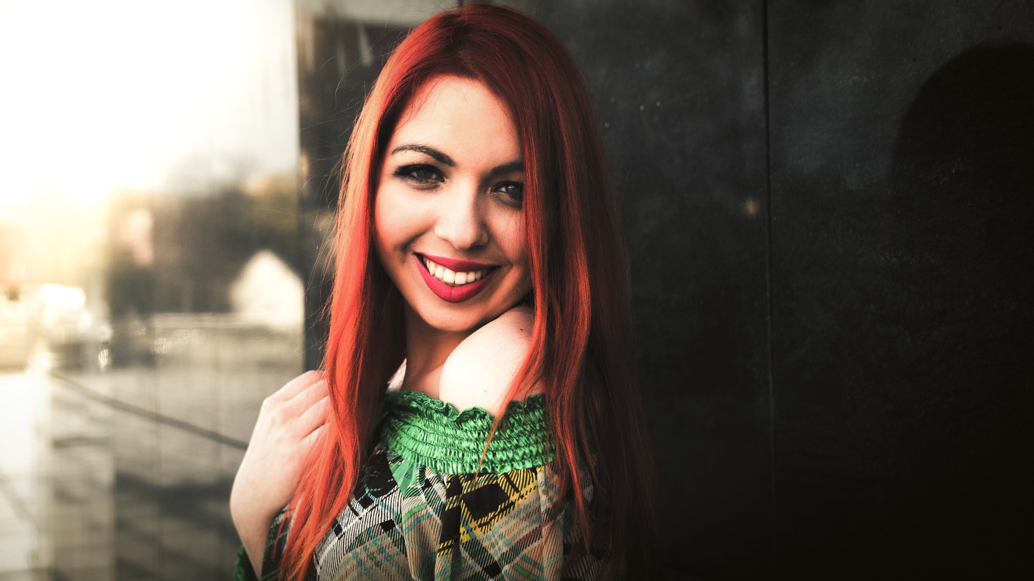 Portret uśmiechniętej kobiety z długimi, rudymi włosami, w kraciastej bluzce z zieloną, marszczoną górą, na rozmytym tle miejskim z elementami architektury i refleksami światła.