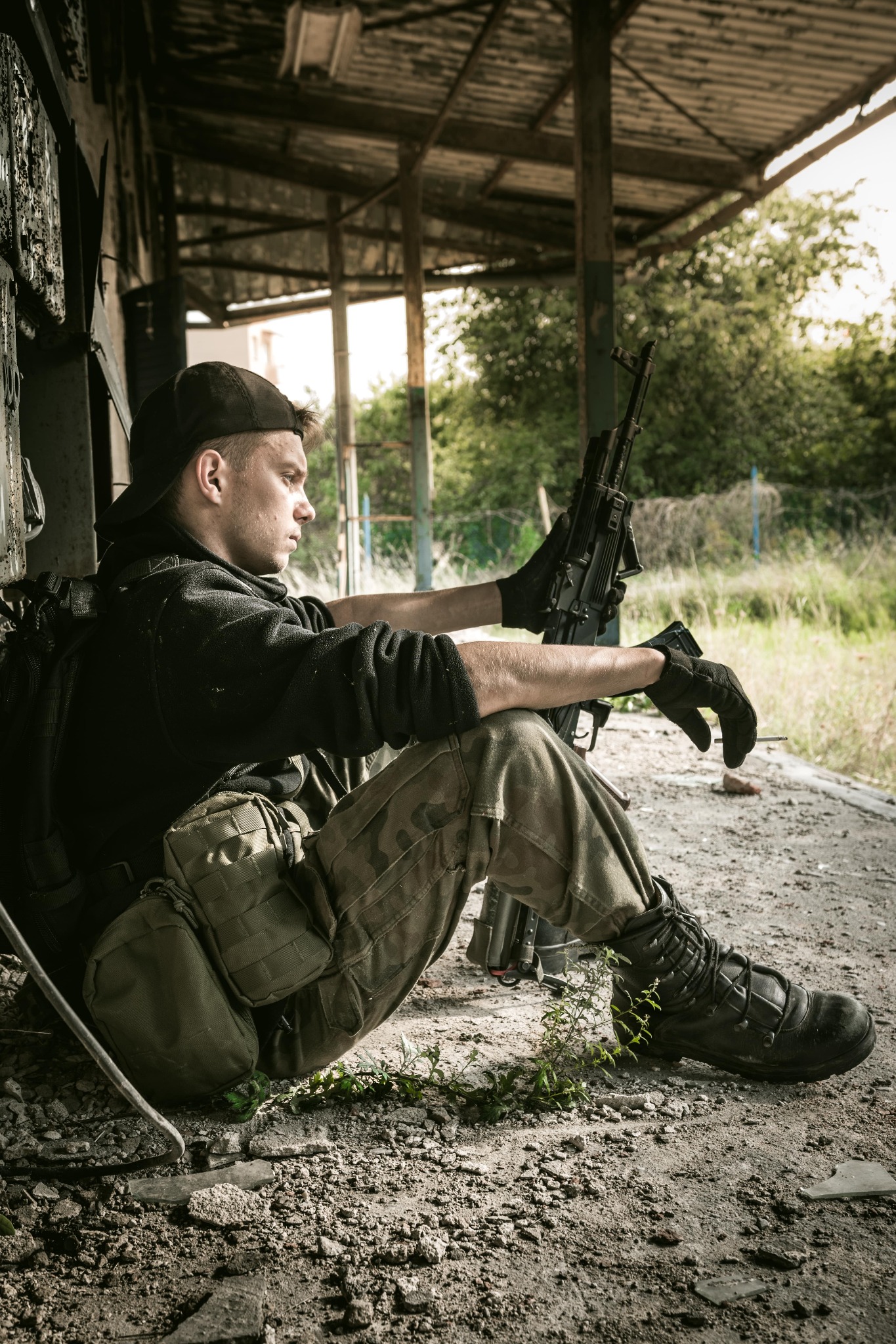 Młody mężczyzna w czarnej bluzie i czapce, w kamuflażowych spodniach i taktycznych butach, siedzi w opuszczonym budynku z karabinem w rękach, spoglądając w dal.