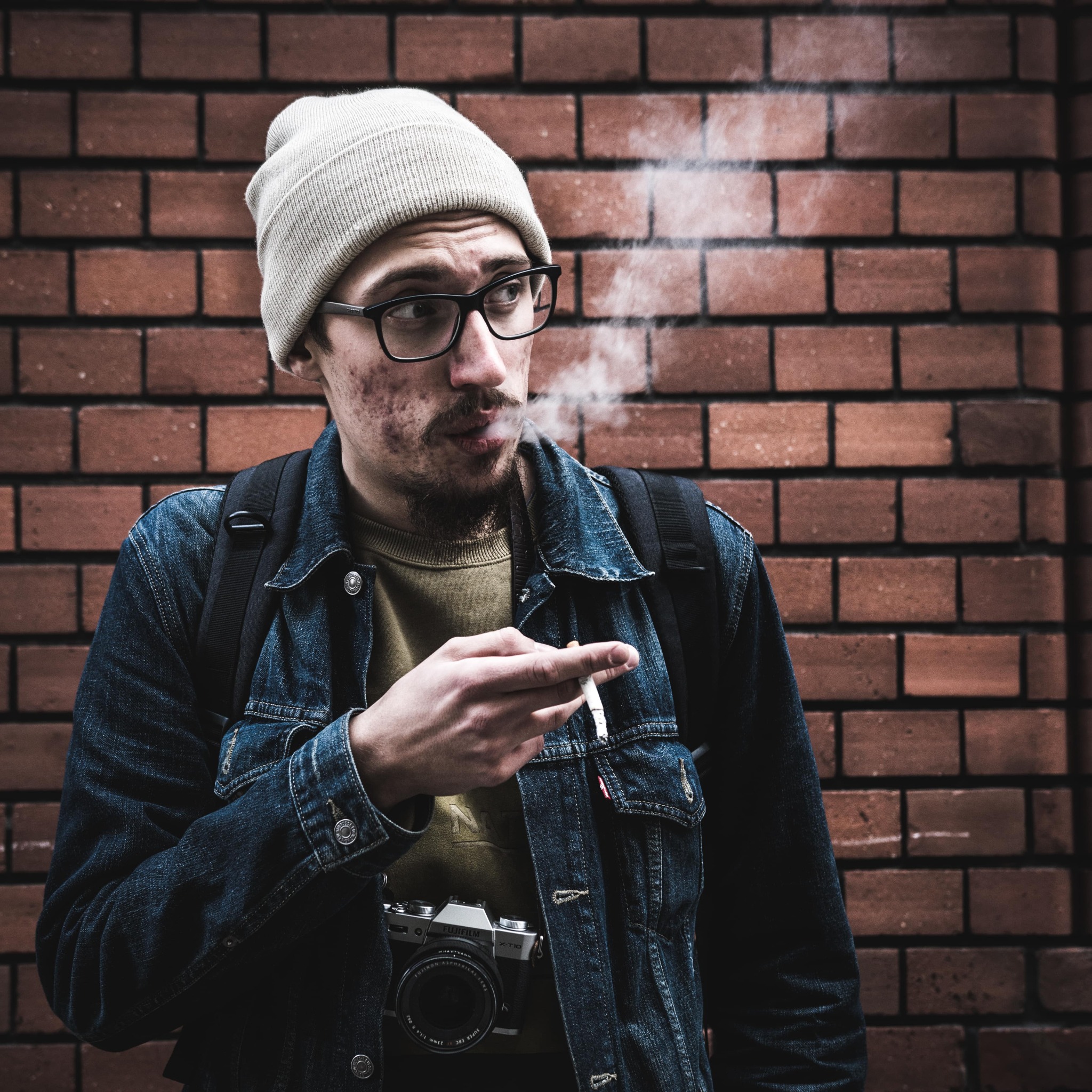 Mężczyzna w czapce i okularach, z aparatem Fujifilm X-T10 na szyi, pali papierosa na tle ceglanej ściany.