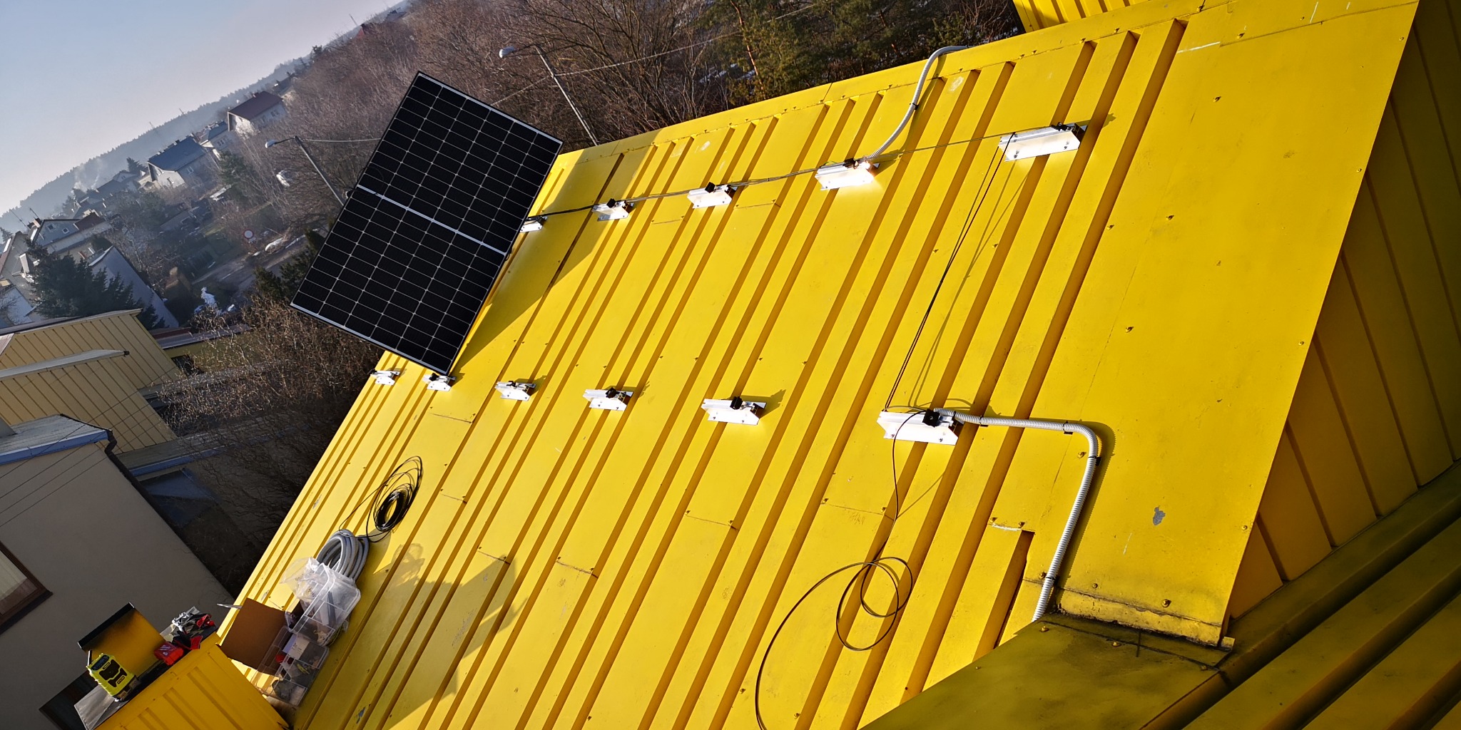 Żółty blaszany dach z zamontowanym pojedynczym panelem fotowoltaicznym i uchwytami pod kolejne, widoczna instalacja elektryczna w peszlu, skrzynka z narzędziami na dachu w rogu.