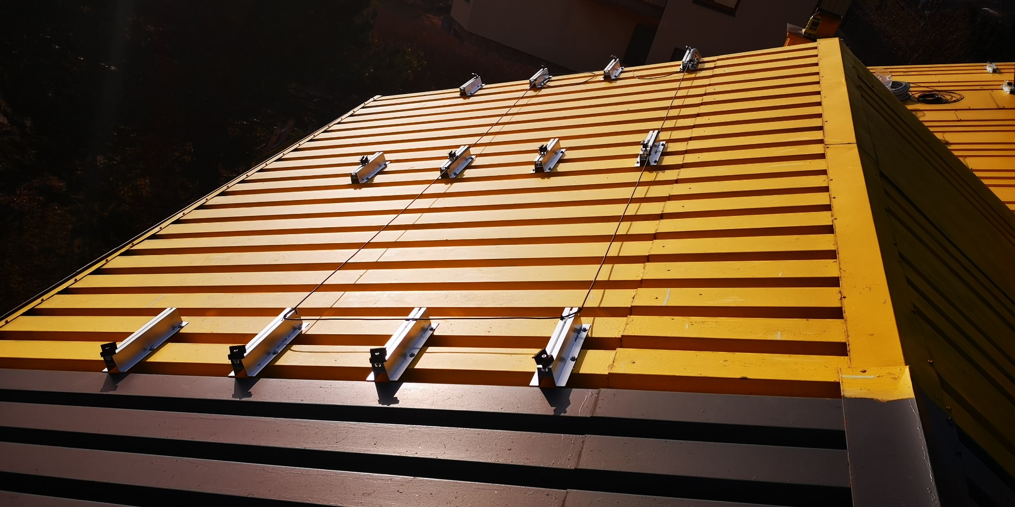 Żółto-brązowy dach z blachy trapezowej z zamontowanymi uchwytami do instalacji odgromowej, widoczne przewody uziemiające.