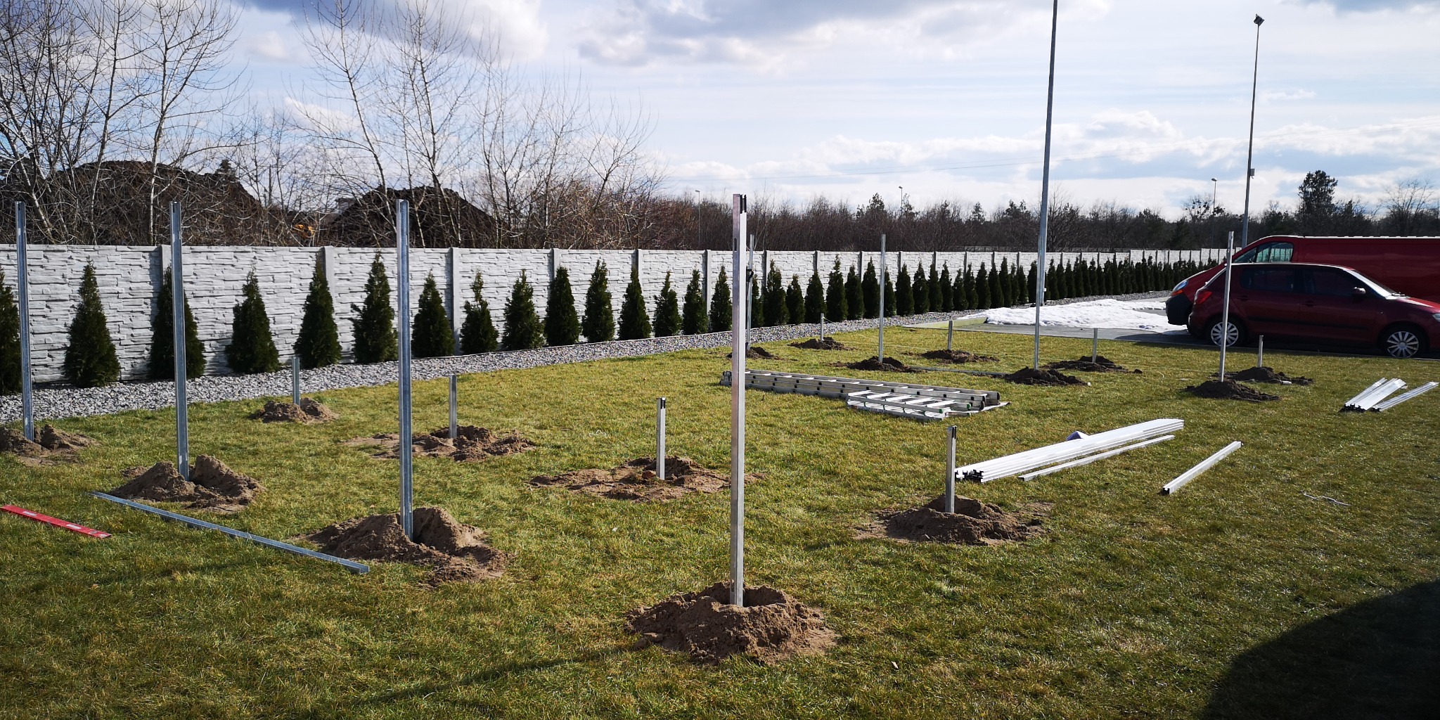 Przygotowanie terenu pod konstrukcję, prawdopodobnie fotowoltaiczną: wbite metalowe słupki w ziemię, rozsypana ziemia, drabina i profile na trawie, w tle ogrodzenie z tujami i zaparkowane samochody.