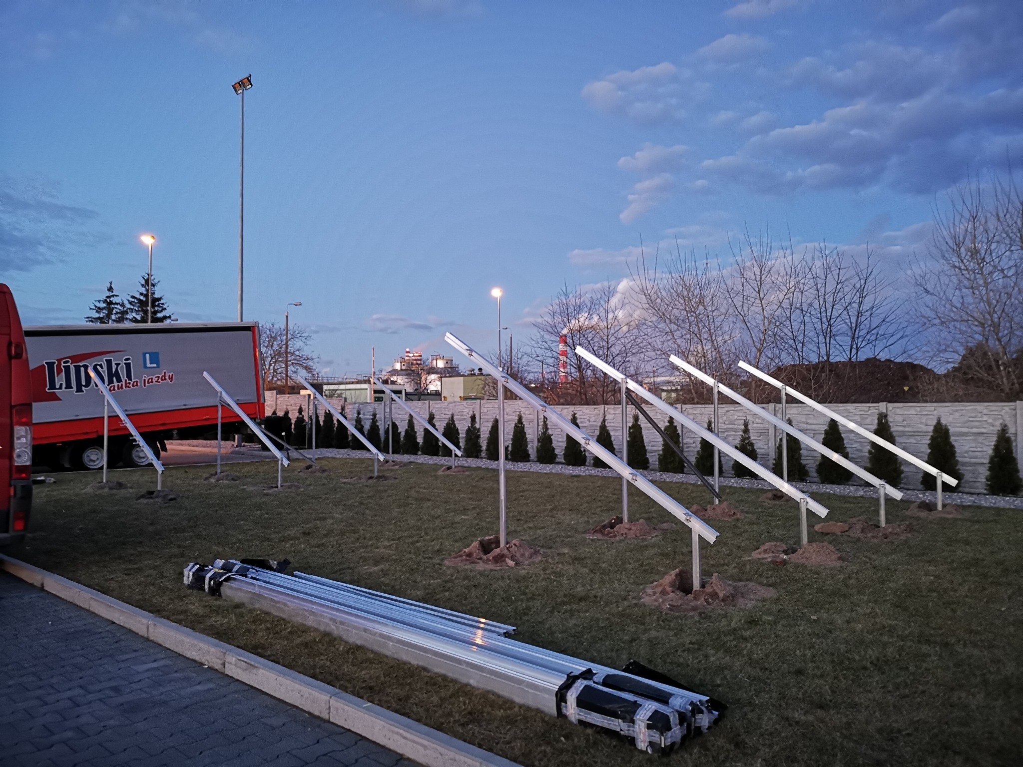 Montaż konstrukcji wsporczej paneli słonecznych na trawniku, widoczny samochód dostawczy firmy Lipski L w tle, wieczorne niebo.