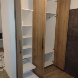 Otwarta szafa wnękowa z drewnianymi drzwiami i białymi półkami oraz wysuwanymi szufladami, widoczne zawiasy i podłoga z paneli imitujących drewno.