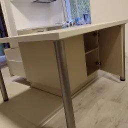 Fragment wyspy kuchennej z otwartą szafką, widoczne metalowe nogi i jasne fronty szafek, w tle okno i zabudowa kuchenna.