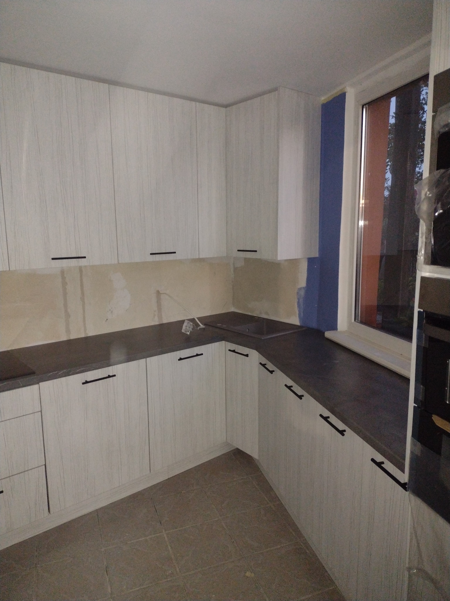 Kątowe ujęcie kuchni w trakcie montażu, widoczne białe szafki z czarnymi uchwytami, szary blat, fragment ściany z widocznymi śladami po pracach wykończeniowych oraz okno.