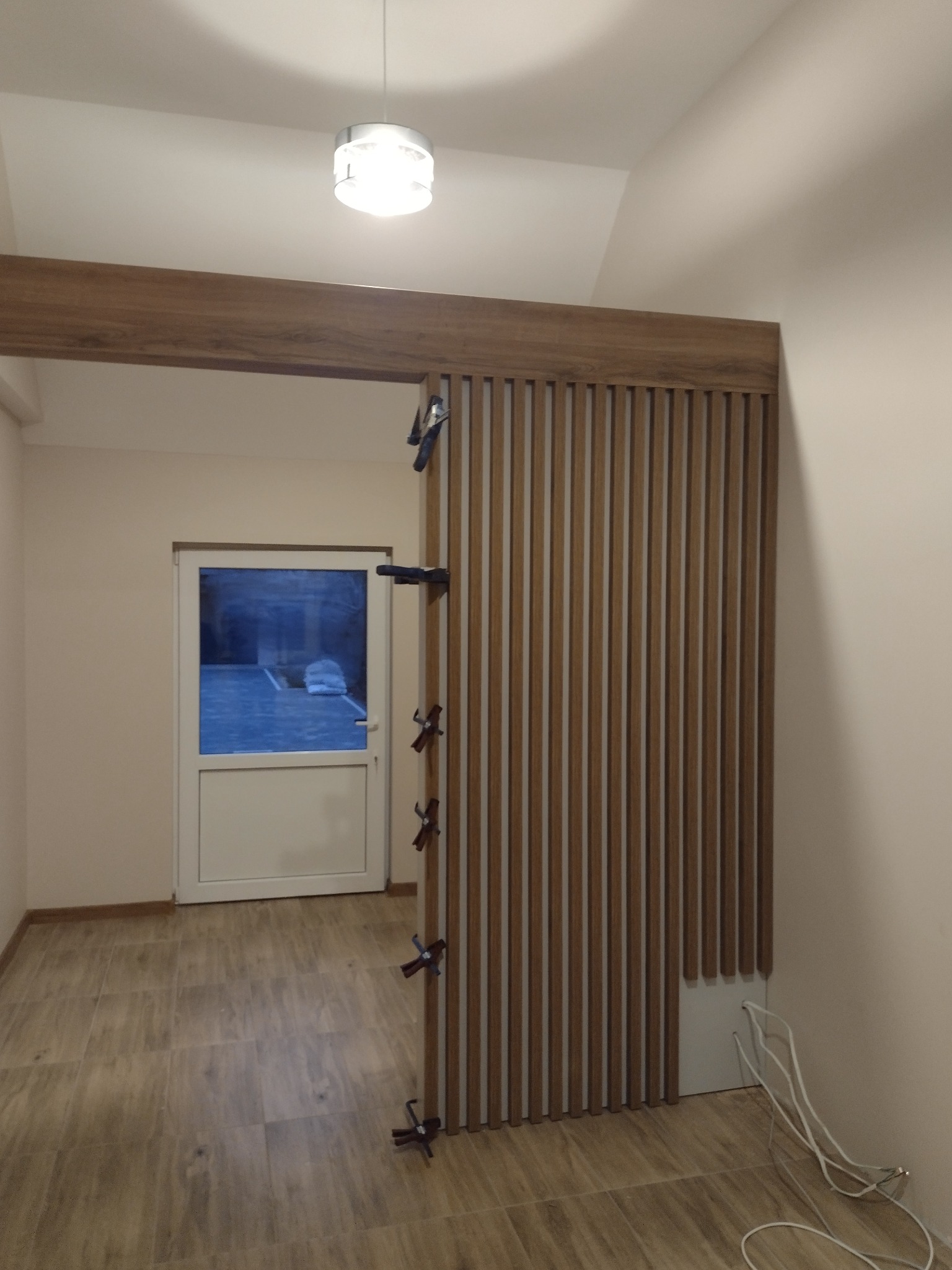 Montaż drewnianej ścianki działowej z lameli, tymczasowo przymocowanej klamrami stolarskimi, widok na podłogę z płytek imitujących drewno i drzwi wejściowe.