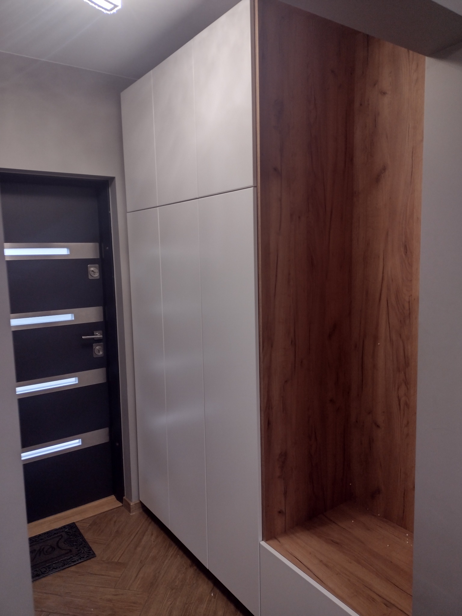 Wysoka szafa wnękowa z białymi frontami i otwartą wnęką z drewnianym wykończeniem, umieszczona w przedpokoju obok drzwi wejściowych z metalowymi elementami dekoracyjnymi.