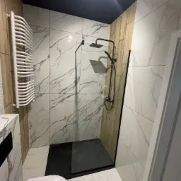 Nowoczesna łazienka z czarną armaturą, kabiną prysznicową typu walk-in z czarnym brodzikiem, grzejnikiem drabinkowym i płytkami imitującymi marmur na ścianach oraz ciemnym sufitem.