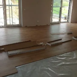 Wnętrze pokoju z częściowo ułożoną podłogą z paneli imitujących drewno, kartony po panelach porozrzucane na podłodze, folia ochronna na podłożu, duże okna balkonowe w tle.