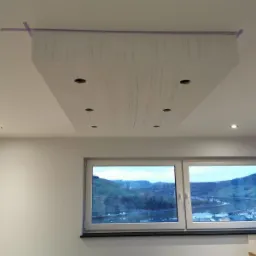 Podwieszany sufit z płyty karton gips z wyciętymi otworami na oświetlenie punktowe, zabezpieczony taśmą malarską na styku ze standardowym sufitem, widok z dołu na okno z widokiem na dolinę.