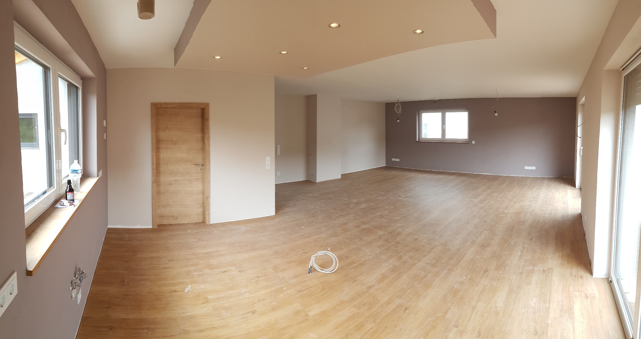 Panoramiczne ujęcie przestronnego wnętrza po remoncie: świeżo pomalowane ściany w odcieniach beżu i brązu, jasna drewniana podłoga, zamontowane oświetlenie sufitowe, drewniane drzwi wewnętrzne...