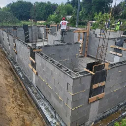 Wznoszenie ścian fundamentowych z bloczków betonowych, z widocznym zbrojeniem i izolacją przeciwwilgociową, pracownik w czerwonej czapce i koszulce z numerem 10 na górze konstrukcji, drugi...