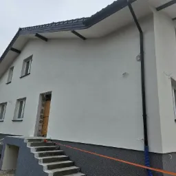 Nowoczesny dom murowany z ocieploną elewacją w jasnych barwach, widoczne schody wejściowe i częściowo wykończony fundament z izolacją.
