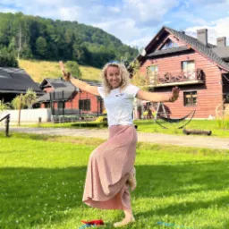 Uśmiechnięta animatorka balansuje na linie slackline na trawie przed drewnianymi domkami w górach, w tle zielone wzgórza i błękitne niebo z chmurami.