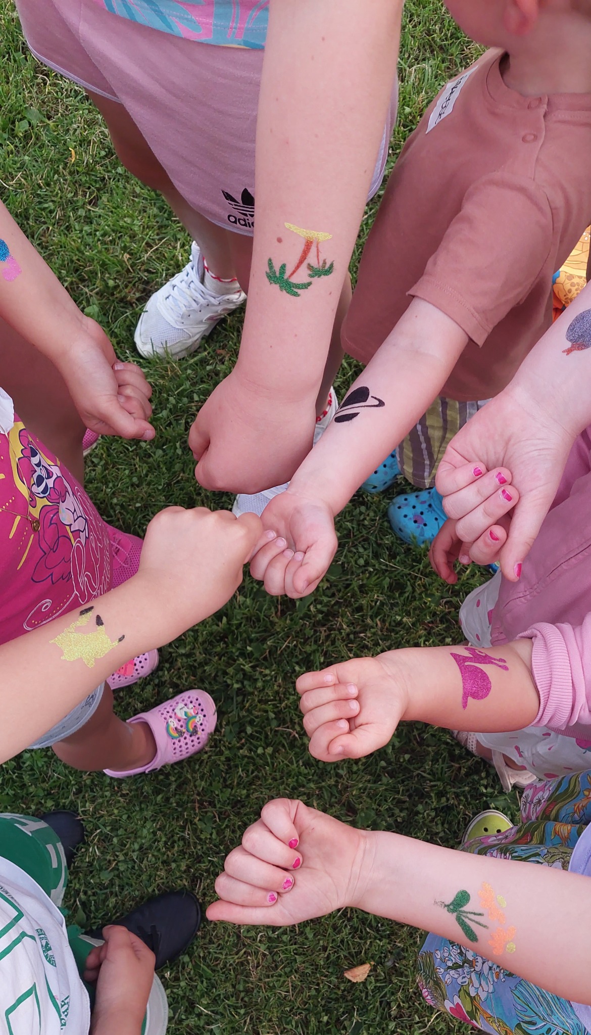 Dzieci na trawie z brokatowymi tatuażami na ramionach, widok z góry, ukazujący różnorodne wzory, takie jak palma, planeta, królik i inne abstrakcyjne motywy.