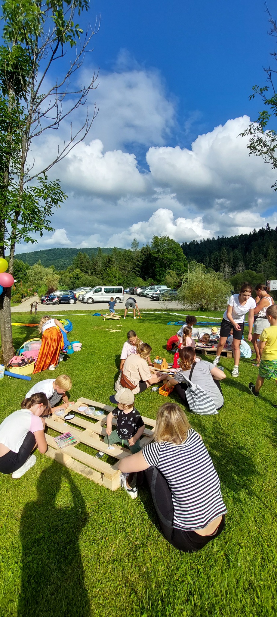 Animatorzy prowadzą warsztaty plastyczne dla dzieci na świeżym powietrzu, wykorzystując drewniane palety jako stoły, w tle zielona trawa, drzewa i góry.