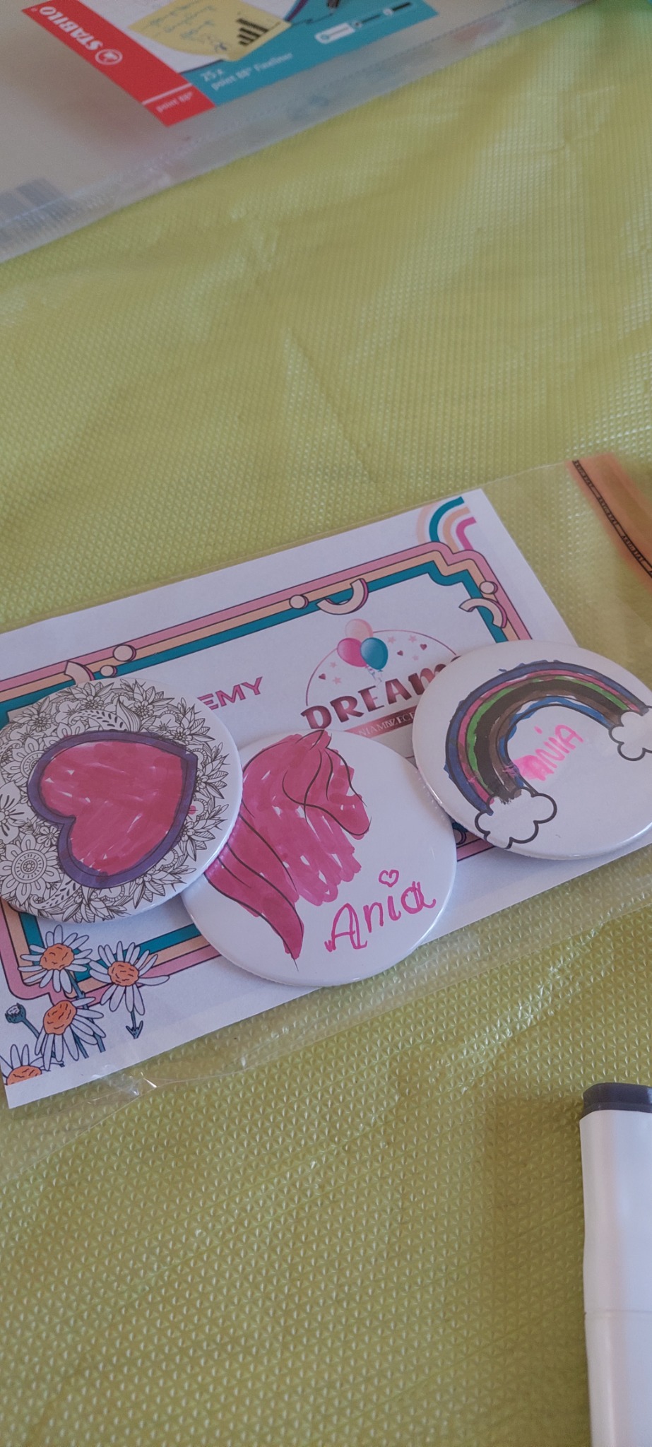Trzy ręcznie robione przypinki z imieniem 'Ania' i rysunkami serca, jednorożca oraz tęczy, leżące na kartce z motywem kwiatów i logo 'Dream Academy'.