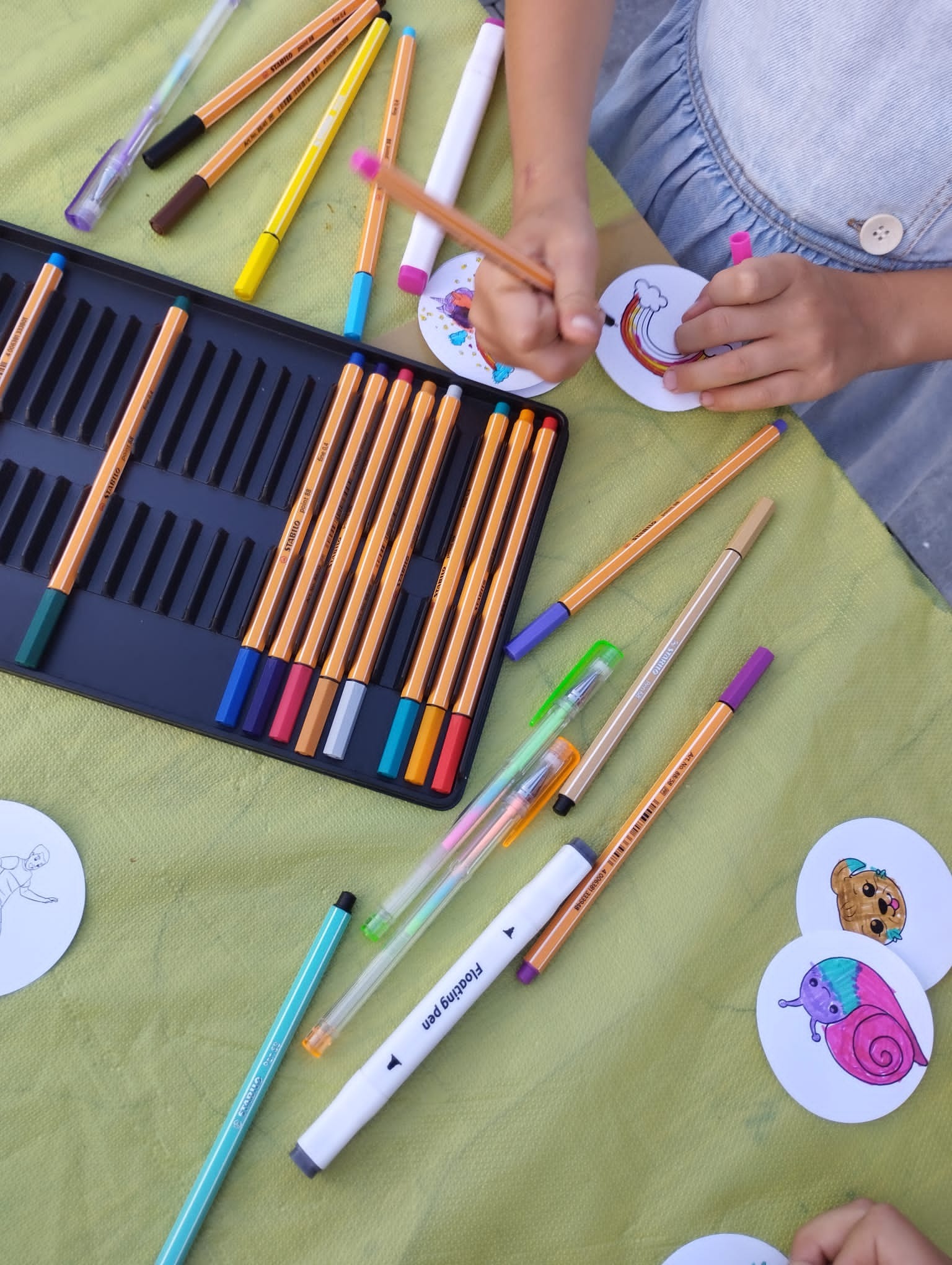 Dziecko koloruje obrazek tęczy na okrągłej kartce, używając cienkopisów Stabilo, z otwartym pudełkiem pełnym kolorowych pisaków, na zielonym tle.