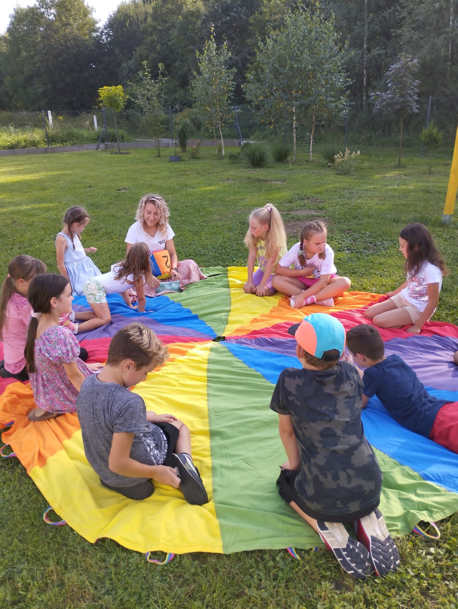 Grupa dzieci i dorosły siedzą na trawie na kolorowej chuście animacyjnej podczas zabawy na świeżym powietrzu.