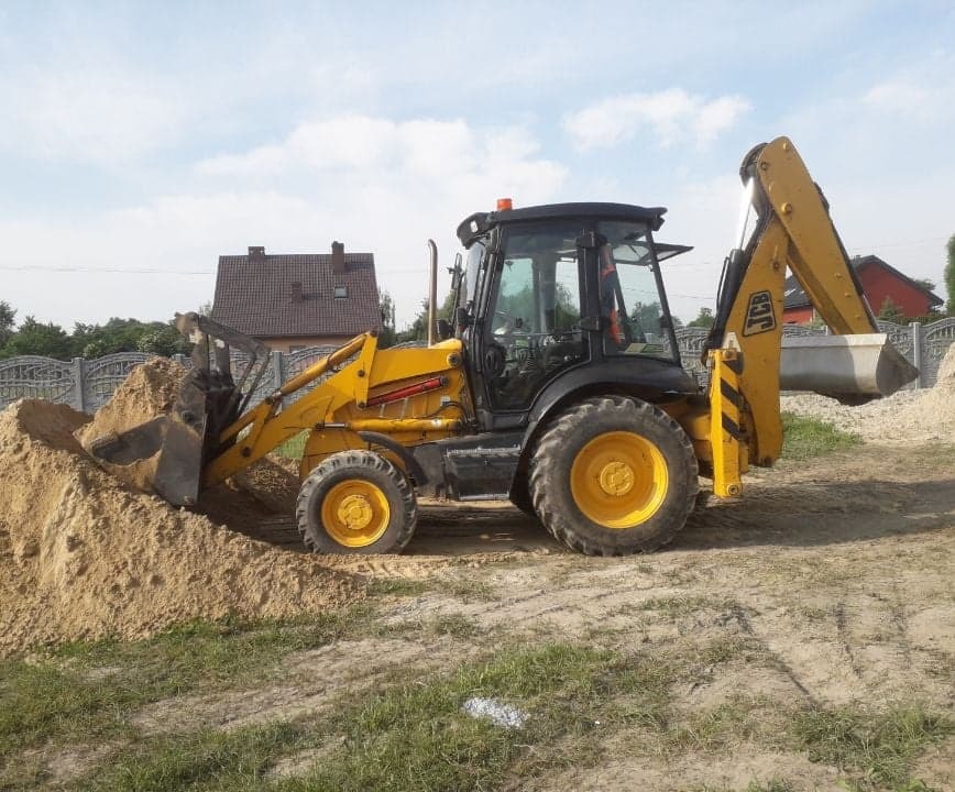 Żółto-czarna koparko-ładowarka JCB nabiera piasek z pryzmy na placu budowy, w tle ogrodzenie i dom.