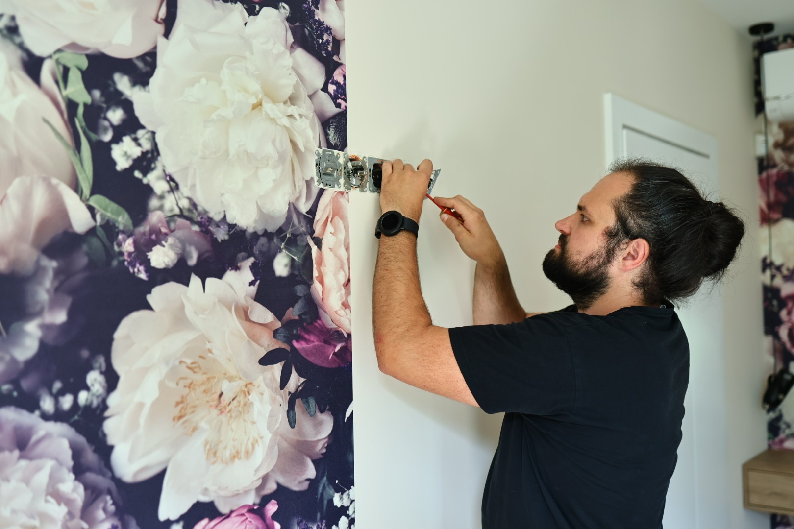 Elektryk z brodą, w czarnej koszulce, montuje gniazdka na ścianie z tapetą w kwiaty, używając czerwonego śrubokręta. Widoczny zegarek na nadgarstku.