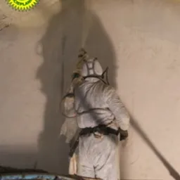 Pracownik w kombinezonie ochronnym natryskuje pianę izolacyjną na ścianę poddasza, widoczny sprzęt do natrysku i częściowe pokrycie powierzchni pianą.