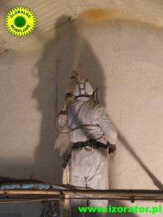 Pracownik w kombinezonie ochronnym natryskuje pianę izolacyjną na ścianę poddasza, widoczny sprzęt do natrysku i częściowe pokrycie powierzchni pianą.