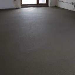 Gładka, szara wylewka w jasnym pomieszczeniu z dużym oknem balkonowym i niedokończonymi ścianami.