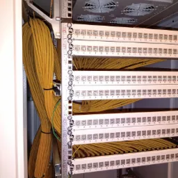 Panel krosowniczy z podłączonymi kablami sieciowymi w szafie rack. Kable uporządkowane, oznaczone numerycznie. Widoczny wentylator w górnej części szafy.