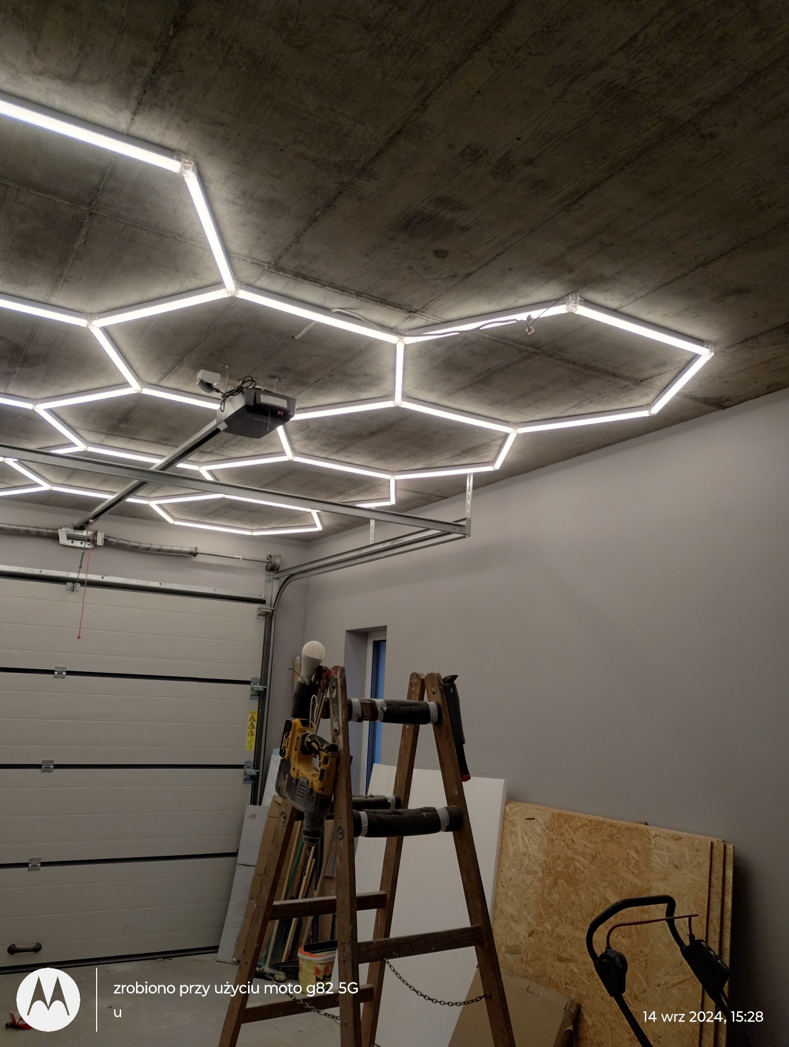 Nowoczesne oświetlenie garażu z heksagonalnymi lampami LED na surowym betonowym suficie. Drabina z narzędziami w tle. Brama garażowa i okno.