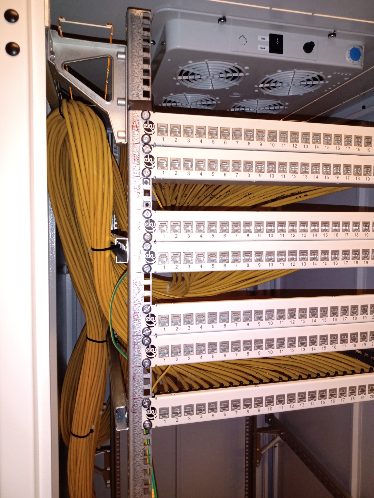 Panel krosowniczy z podłączonymi kablami sieciowymi w szafie rack. Kable uporządkowane, oznaczone numerycznie. Widoczny wentylator w górnej części szafy.