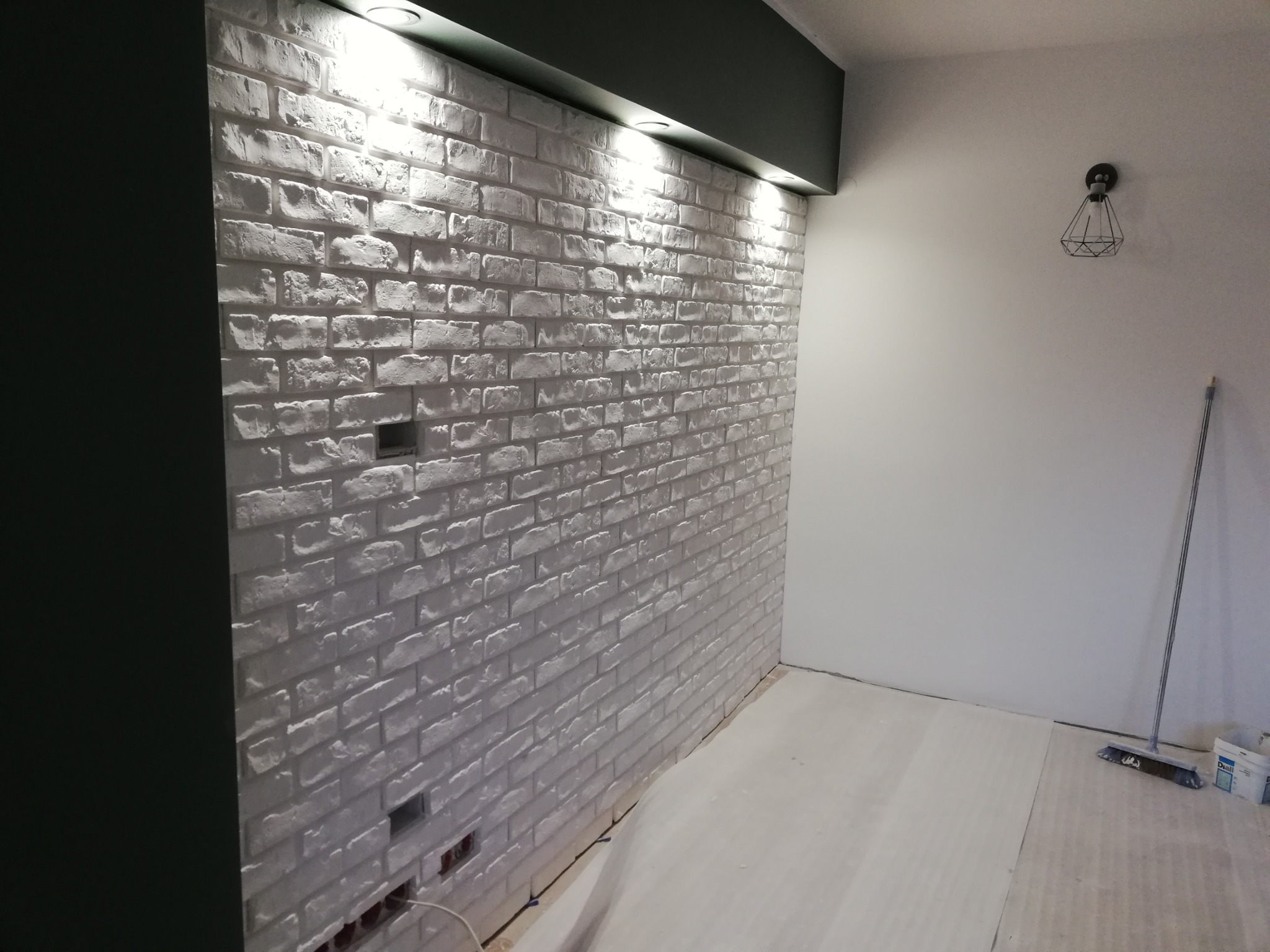 Ściana wykończona białą cegłą dekoracyjną z widocznymi puszkami elektrycznymi, oświetlona reflektorami sufitowymi, z fragmentem ciemnej zabudowy i lampą wiszącą w tle.