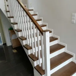 Nowoczesne schody z drewnianymi stopniami i białymi balustradami, widoczne niedokończone wykończenie ściany w tle.