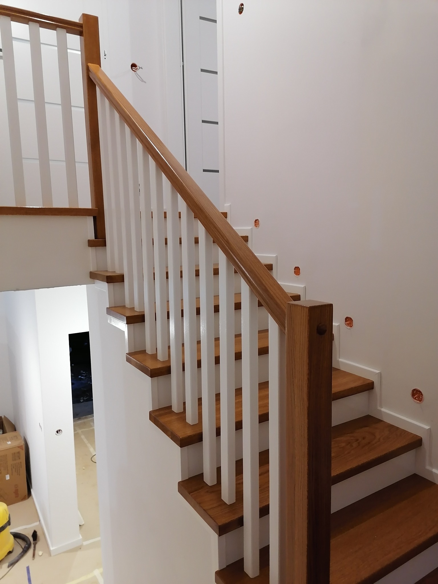 Drewniane schody z białymi balustradami i drewnianą poręczą, widoczne puszki elektryczne w ścianie, pomieszczenie w trakcie wykańczania.
