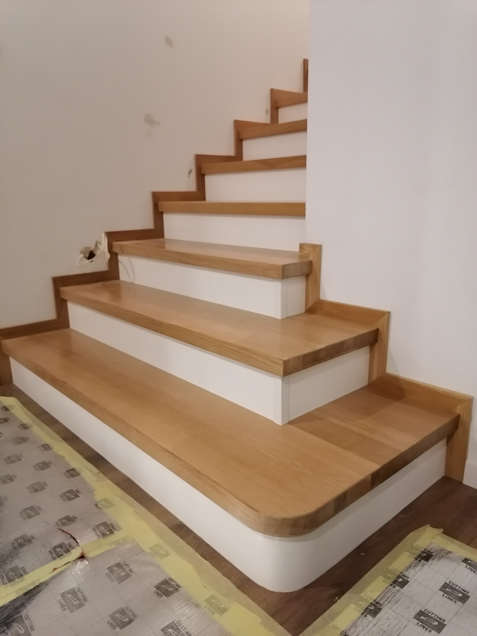 Nowoczesne schody z drewna dębowego z białymi podstopnicami, zabezpieczone folią ochronną podczas montażu, widoczne niedoskonałości ściany w tle.
