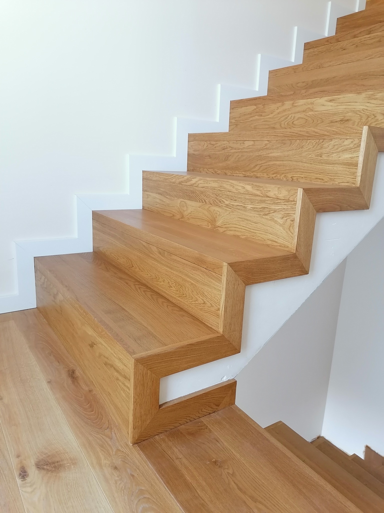 Nowoczesne schody drewniane z jasnego drewna dębowego, z minimalistycznym wykończeniem białych ścian. Charakterystyczne, geometryczne stopnie bez podstopnic.