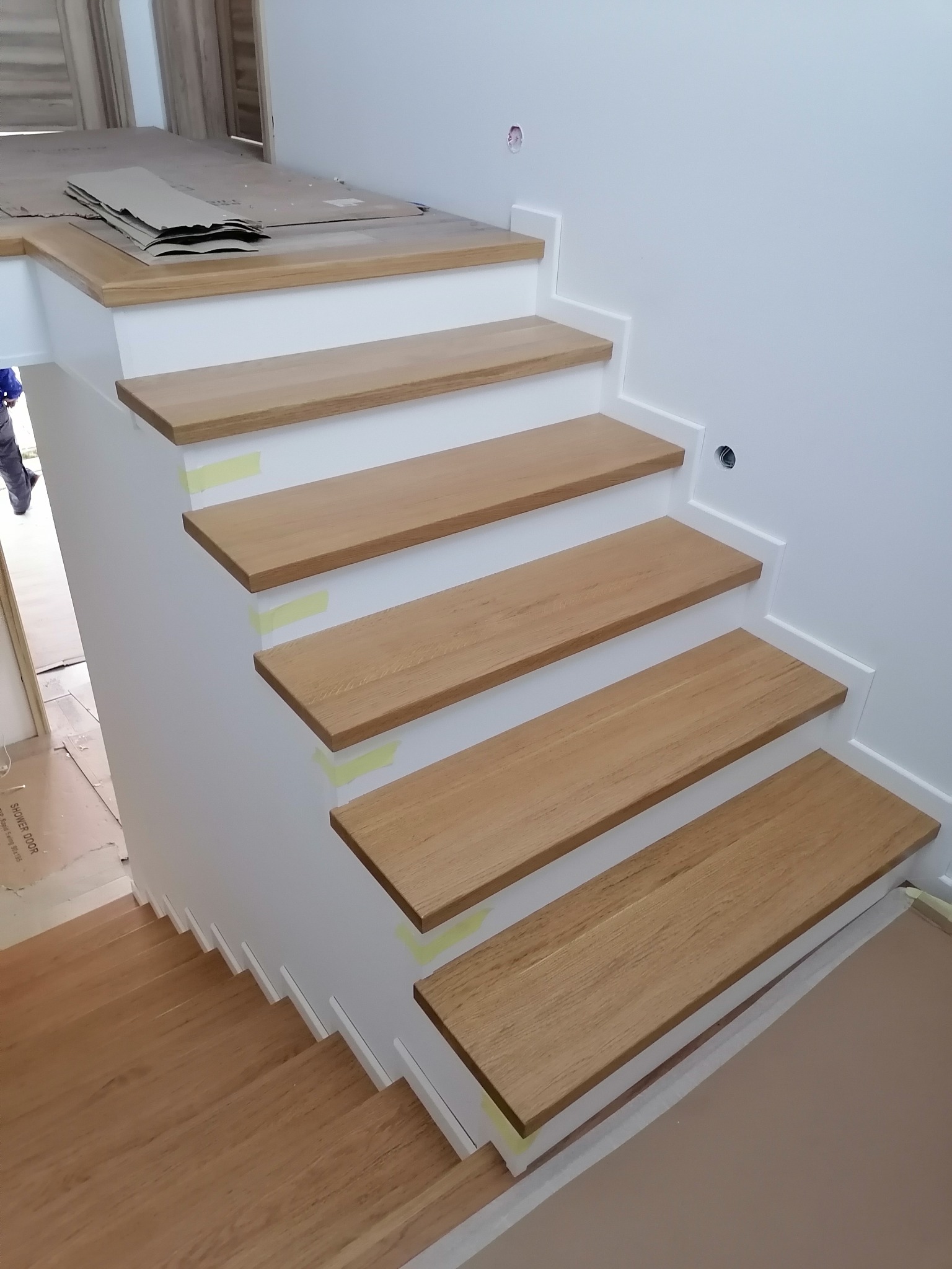 Białe schody z drewnianymi stopniami w trakcie montażu, zabezpieczone taśmą malarską, widoczne przygotowania do dalszych prac wykończeniowych.