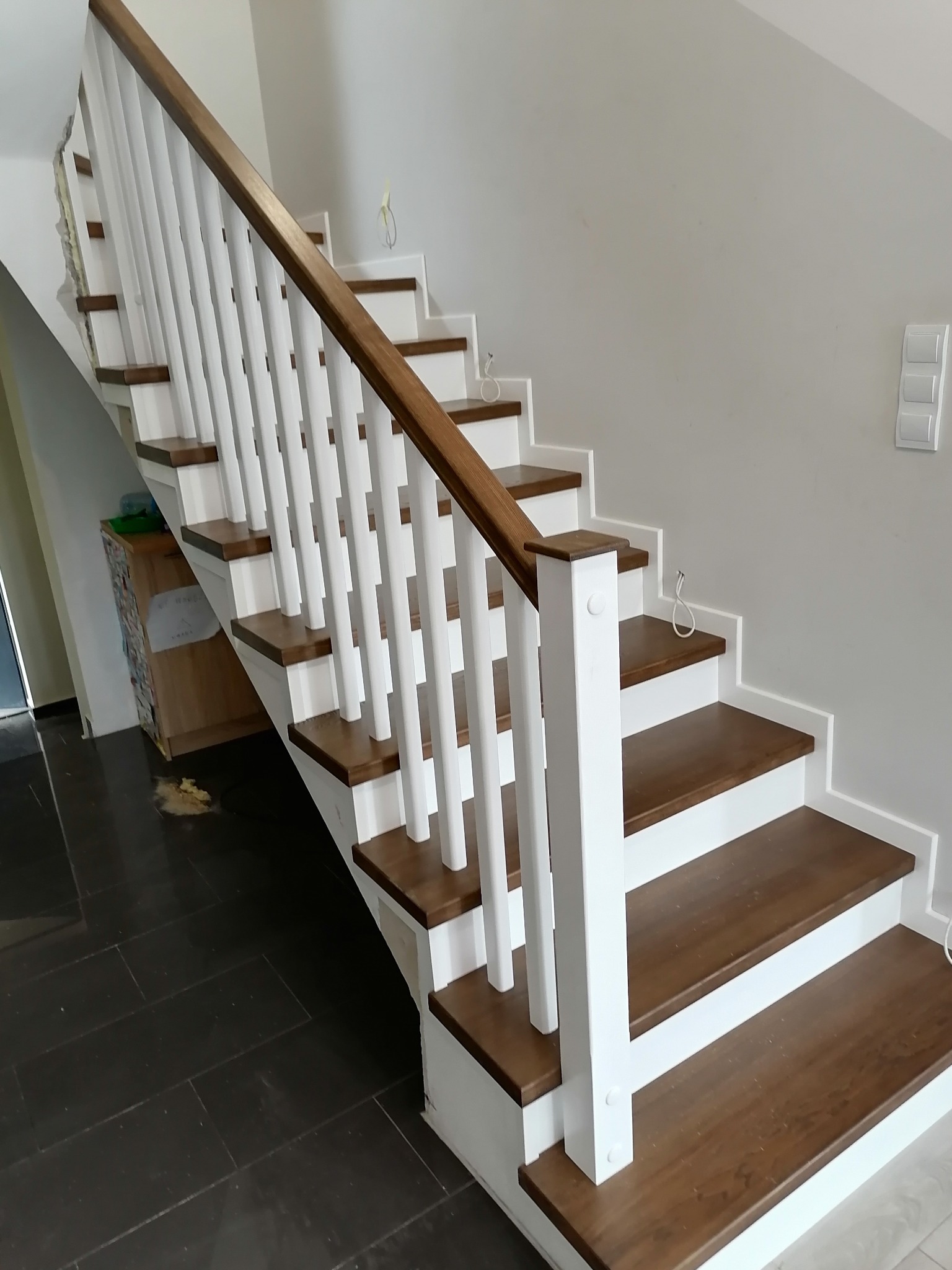 Nowoczesne schody z drewnianymi stopniami i białymi balustradami, widoczne niedokończone wykończenie ściany w tle.